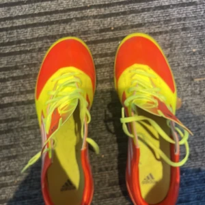 Adidas fotbollsskor i neon och rött - Säljer ett par Adidas fotbollsskor i riktigt snygg neon-gul och röd färg med vita ränder på sidorna. Skorna har snörning och dobbar för gräsplan. Perfekta för dig som vill sticka ut på planen och få bra grepp när du spelar. Sköna dojor att lirka runt i under spontanfotboll