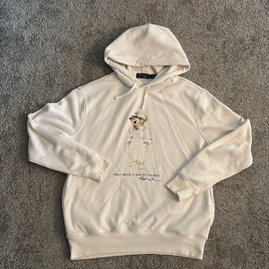 Vit hoodie Polo Bear från Ralph Lauren - Vit hoodie från Polo Ralph Lauren med ikoniskt Polo Bear-tryck på bröstet. Tröjan har huva med snörning och ribbade muddar. Materialet är mjuk bomull, perfekt för chill dagar. Klassisk och stilren design med coolt motiv framtill. Lite sliten på märket men inge mer än de.
