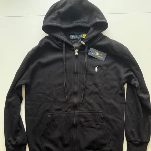 Polo Ralph Lauren Zip Hoodie Svart - Svart hoodie från Polo Ralph Lauren med dragkedja framtill, huva med snörning och klassisk broderad logga på bröstet. Tröjan är helt ny och har inga spår av skador eller användning. Den är i storleken M men modellen är mer som S skulle jag säga.