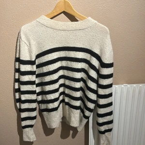 Randig stickad tröja i svart och vitt/beige - Mysig stickad tröja med breda svarta och vita ränder. Tröjan har lång ärm, ribbade muddar och en avslappnad passform. Perfekt för kyliga dagar!