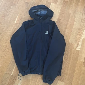 Svart vindjacka från Haglöfs dam M - Svart vindjacka från Haglöfs i dammodell med huva och meshfoder. Jackan har dragkedja framtill, elastiska muddar och justerbar nederkant. Perfekt för blåsiga dagar och outdoor-äventyr. Snygg och funktionell design med diskret logga på bröstet.