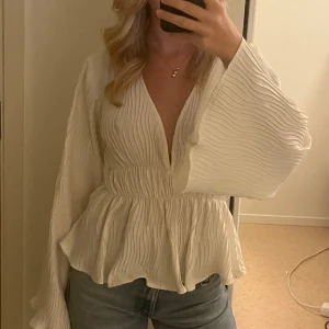 Vit plisserad blus med v-ringning - Säljer en vit blus med plisserat tyg och djup v-ringning. Blusen har vida, långa ärmar och markerad midja med resår. Tyget är lätt och ger en flowy känsla. Perfekt att styla med jeans eller kjol för en trendig look.