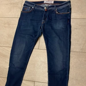 Jacob Cohën mörkblå jeansbyxor - Snygga mörkblå jeansbyxor från Jacob Cohën med orange kontrastsömmar och klassisk femficksdesign. Byxorna har raka ben och är tillverkade i mjukt jeansmaterial. Perfekta för dig som gillar stilrena och exklusiva jeans med unika detaljer.