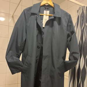 Mörkblå trenchcoat från Lindbergh - Stilren mörkblå trenchcoat från Lindbergh med klassisk krage och knäppning framtill. Jackan har raka linjer, två sidofickor och är helfodrad. Perfekt för dig som vill ha en clean och tidlös look. Aldrig använd. Pris lapp sitter kvar nypris 1800