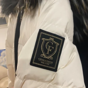 Vit pufferjacka från Chiara Forthi(ÄKTA) - Snygg vit pufferjacka från Chiara Forthi med svart fuskpäls på huvan och svarta detaljer vid dragkedjorna. Jackan har quiltad design, två fickor med dragkedja och en stor logotyp på ärmen. Perfekt för kalla dagar när du vill vara både varm och stilren. Den har lite smink fläckar vid kragen som jag gärna vill informera om. Priset kan vi ta privat. 