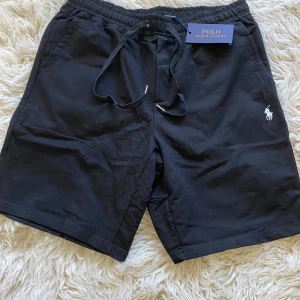 Svarta shorts från Polo Ralph Lauren - Svarta shorts från Polo Ralph Lauren med klassisk broderad logga i vitt på benet. De har elastisk midja med snörning och är tillverkade i mjuk bomull. Perfekta för en chill och stilren look.