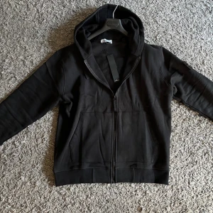 Svart hoodie från Stone Island - Svart hoodie från Stone Island med dragkedja framtill, huva med snörning och klassisk logotyp-patch på ärmen.  