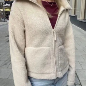 Beige teddyjacka med dragkedja - Mysig beige teddyjacka med dragkedja framtill och stora fickor. Jackan har en hög krage och är tillverkad i mjukt fleece-liknande material. Perfekt för kyliga dagar och enkel att matcha med jeans eller andra plagg.