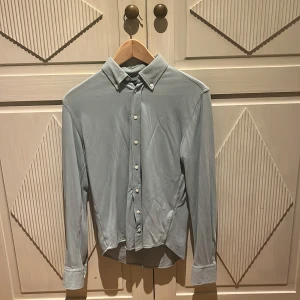 Ljusgrön Hackett skjorta - Snygg ljusgrön skjorta från Hackett med klassisk button down-krage och broderad logga på bröstet. Skjortan har långa ärmar, vita knappar och är tillverkad i mjuk bomull. Perfekt för en clean och stilren look.