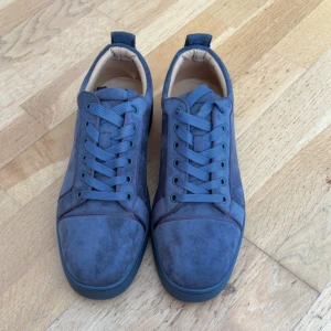 Gråa mockasneakers från Louboutin - Snygga gråa sneakers i mocka från Christian Louboutin med klassisk röd sula och rund tå. Skorna har ton-i-ton snörning och diskreta sömdetaljer. Insidan är ljusbeige och sulan har den ikoniska Louboutin-signaturen.