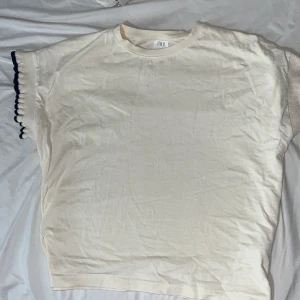Vit t-shirt med volangärm från Zara - Vit t-shirt från Zara i storlek 152. T-shirten har korta ärmar med volangdetalj och mörkblå kant. Rund halsringning och mjukt bomullsmaterial gör den skön att bära. Perfekt för dig som gillar stilrena plagg med en twist. Ny utan prislapp