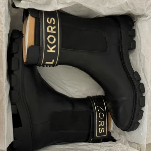 Svarta boots från Michael Kors - Stilrena svarta boots från Michael Kors i skinn med chunky sula och elastiska paneler på sidorna. Runt skaftet finns en bred rem med guldiga detaljer och tydlig logga. Perfekta för dig som vill ha en edgy och lyxig look.