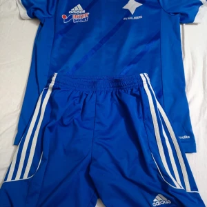 Blå IFK Hallsberg fotbollströja Adidas - Säljer en blå fotbollströja från Adidas med IFK Hallsberg-tryck och Intersport-logga. Tröjan har korta ärmar och klassiska vita Adidas-detaljer. Materialet är lätt och andas, perfekt för träning eller match.
