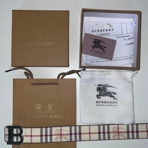 Burberry vändbart bälte med B-spänne - Snyggt vändbart bälte från Burberry med klassiskt rutigt mönster i beige, svart och rött på ena sidan och svart präglat skinn på andra. Stort metallspänne med B-logga. Bältet är 115cm långt, Kommer med originalpåse, box och kvitto. Perfekt accessoar för att lyfta din outfit. Bara till att skriva vid minsta fundering /styleish 