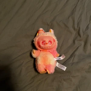 Labubu - Bilderna visar en Pop Mart THE MONSTERS nyckelring/figur i plysch och plast, med rosa och vita färger. Produkten är i mycket fint skick utan synligt slitage. Perfekt som samlarobjekt eller accessoar. Söt design med stora ögon och ett glatt leende.