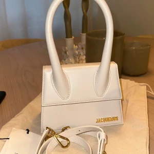 Vit Jacquemus handväska i skinn - Superfin liten handväska från Jacquemus i vit skinn med guldiga detaljer och logga framtill. Väskan har ett stilrent och modernt uttryck med rundade handtag och avtagbar axelrem. Nypris: 7900. Väskan är aldrig använd. Totebag medföljer. 