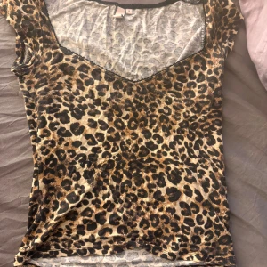 Leopardmönstrad v-ringad topp - Säljer en slim leopardmönstrad topp med v-ringning och korta ärmar. Toppens färger går i beige, brun och svart och materialet är tunt och stretchigt, troligen polyester. Perfekt för dig som vill sticka ut med djurmönster och cool stil.