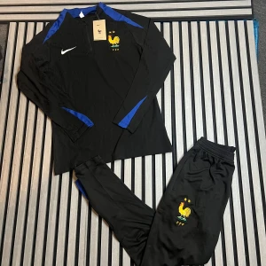 Nike FFF svarta träningsbyxor - Svarta träningsbyxor från Nike med franska landslagets FFF-logga broderad i gult och blått på låret. Byxorna har smal passform, elastisk midja och blå detaljer längs benen. Perfekta för fotbollsträning eller chill.