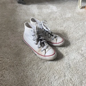 Vita Converse All Star high-tops - Klassiska vita Converse All Star high-tops med röd och blå rand på sulan i storlek 32. Skorna har snörning, rund tå och är tillverkade i textil med gummisula. Perfekta för en cool och avslappnad stil. Min systers gamla som hon nu har växt ur💗 