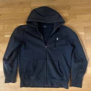 Svart hoodie från Polo Ralph Lauren - Svart hoodie från Polo Ralph Lauren med dragkedja och klassisk vit logga på bröstet. Tröjan har huva med snörning, känguruficka och ribbade muddar. Perfekt för en chill och stilren look.