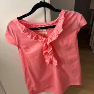 Rosa blus med volangdetalj - Söt rosa blus med korta ärmar och volang längs v-ringningen. Perfekt för dig som gillar färg och vill ha något som sticker ut lite extra. Blusen har en mjuk och lätt känsla, vilket gör den skön att bära under varmare dagar.