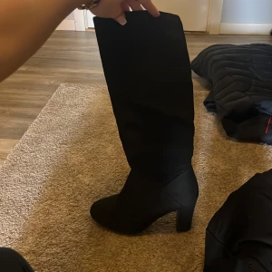 Svarta höga stövlar med klack, strl 38 - Säljer ett par svarta höga stövlar med bred klack och spetsig tå. Skorna har dragkedja på insidan och är tillverkade i ett mockaliknande material. Perfekta för att lyfta din outfit under höst och vinter.