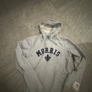 Grå hoodie från Morris med tryck - Grå hoodie från Morris med mörkblå tryck på bröstet och en klassisk huva med snörning. Tröjan har lång ärm, ribbade muddar och en stor ficka framtill. Perfekt för chill dagar och har en vintage-vibe. Materialet är mjuk bomull. Priset kan diskuteras.
