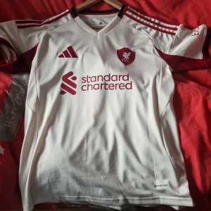 Liverpool vit bortatröja Frimpong 30 - Liverpool FC:s vita bortatröja från Adidas med röda detaljer, klubbmärke och sponsortryck framtill. Frimpong 30 och Premier League-tryck på ryggen. Klassisk V-ringning, korta ärmar och tillverkad i lätt funktionsmaterial (AEROREADY).funkar också om man har s