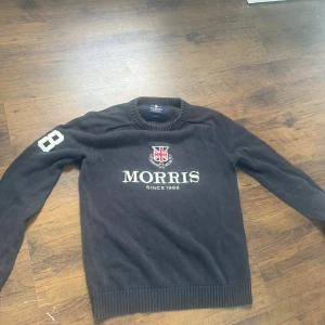 Svart stickad tröja Morris S - Svart stickad tröja från Morris i storlek Small. Tröjan har ett broderat Morris-emblem med brittisk flagga på bröstet och siffran 8 på vänster ärm. Rund hals och ribbade muddar vid ärmslut och nederkant. Perfekt för dig som gillar klassisk stil.
