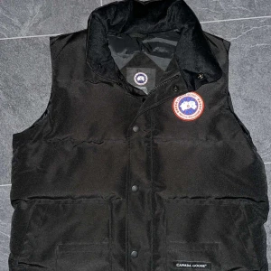 Svart dunväst från Canada Goose - Svart dunväst från Canada Goose med klassisk logga på bröstet och diskret varumärkeslapp på fickan. Västen har hög krage, tryckknappar framtill och två stora fickor. Perfekt för lager-på-lager och riktigt snygg till streetwear-stilen.