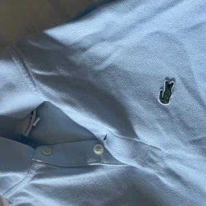 Ljusblå pikétröja från Lacoste - Säljer en klassisk ljusblå pikétröja från Lacoste med den ikoniska gröna krokodilloggan på bröstet. Tröjan har krage, knäppning med två knappar och är tillverkad i bomullspiké som ger en sportig och fräsch känsla.