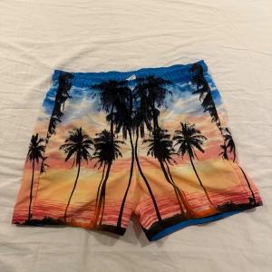Färgglada badshorts från Calliope - Säljer ett par färgglada badshorts från Calliope med tropiskt palmmotiv i blått, orange, rosa och svart. Shortsen har elastisk midja med snörning och innerbyxa i mesh. Perfekta för stranden eller poolen!