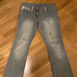 Ljusblå bootcut jeans med strassdetaljer - Säljer ett par ljusblå bootcut jeans med slitningar och coola strassdetaljer på hällor och ena bakfickan. Jeansen har klassisk femficksdesign och är tillverkade i denim. Perfekta för dig som vill sticka ut lite extra.