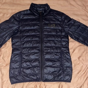 EA7 jacka - Snygg svart pufferjacka från EA7 Emporio Armani med guldfärgat logotryck på bröstet. Jackan har ståkrage, quiltad design och dragkedja framtill med EA7-detalj. Perfekt för dig som vill ha en stilren och modern look under kyliga dagar. Inga skador eller fläckar, inte använt så länge