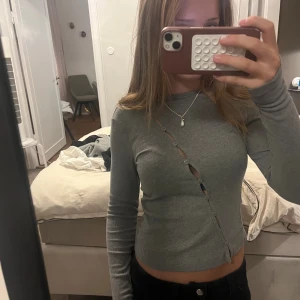 Grå ribbad långärmad tröja med knappar - Grå långärmad tröja från Monki i ribbat material. Tröjan har en unik diagonal knappdetalj framtill som ger en cool twist. Perfekt till jeans eller kjol för en chill look. Materialet är mjukt och stretchigt för extra komfort.