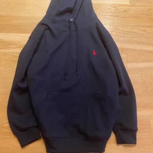 Mörkblå hoodie från Polo Ralph Lauren - Snygg mörkblå hoodie från Polo Ralph Lauren med klassisk röd broderad logga på bröstet. Tröjan har huva med snörning, magficka och ribbade muddar. Tillverkad i mjuk bomull för maximal komfort. Perfekt för en chill och stilren look.