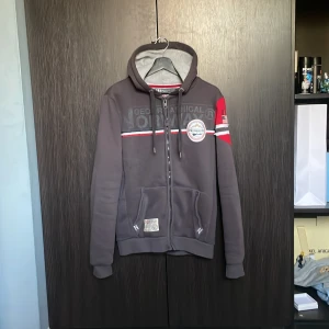 Grå hoodie från Geographical Norway - Säljer en grå hoodie från Geographical Norway med dragkedja, huva och snörning. Tröjan har röda och vita detaljer på ärmen, broderade loggor och två fickor framtill. Perfekt för dig som gillar sportig och avslappnad stil.