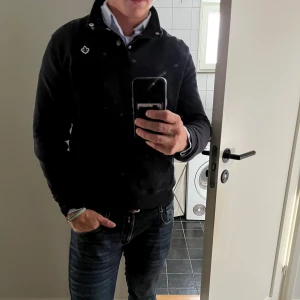 Svart långärmad pikétröja Morris strl M - Svart långärmad pikétröja från Morris med klassisk krage, knappar vid halsen och broderad logga på bröstet. Tröjan har en stilren look och är tillverkad i bomullsmix som känns mjuk mot huden. Perfekt för dig som gillar en clean och tidlös stil.