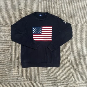 Mörkblå stickad tröja GANT med flagga - Mörkblå stickad tröja från GANT med amerikansk flagga broderad på bröstet och texten 'GANT NH-CT 1949' på ena ärmen. Rund halsringning och ribbade muddar. Perfekt för dig som gillar klassisk preppy stil.
