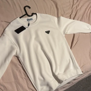 Vit sweatshirt från Prada - Säljer en stilren vit sweatshirt från Prada med klassisk rund hals och svart logotyp-bricka på bröstet. Tröjan har ribbade muddar vid ärmslut och nederkant. Perfekt för dig som gillar minimalistisk och exklusiv stil. Pris kan diskuteras vid snabb affär
