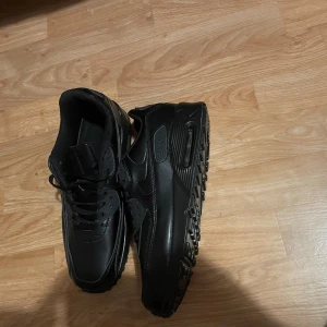 Nike Air Max 90 helsvarta sneakers - Säljer ett par helsvarta Nike Air Max 90 sneakers i storlek 43.5. Skorna har klassisk chunky sula med synlig Air Max-dämpning, snörning och ovandel i skinn och syntet. Perfekta för dig som gillar en clean och stilren look.