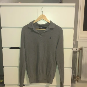 Grå pikétröja från Polo Ralph Lauren - Säljer en klassisk grå pikétröja från Polo Ralph Lauren med broderad marinblå logga på bröstet. Tröjan har lång ärm, krage och knäppning framtill. Tillverkad i mjuk bomull som känns skön mot huden.