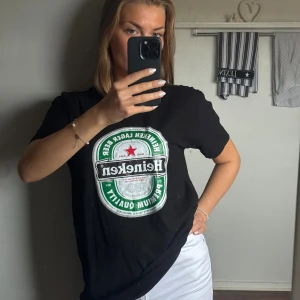 Svart Heineken t-shirt - Svart t-shirt med stort Heineken-tryck på bröstet. Klassisk rund hals och korta ärmar. Materialet är mjuk bomull och passformen är normal🍺Köpte den och har tvättats en gång men aldrig använts