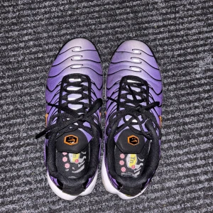 Nike Air Max Plus TN lila och svart - Nike Air Max Plus TN sneakers i lila och svart med orange detaljer och svart snörning. Skorna har en vågig design på ovandelen, synlig Air Max-dämpning i sulan och meshmaterial för extra komfort. Swoosh-logga i orange på sidan och Tn-detaljer på häl och sula.