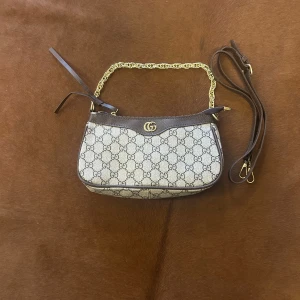 Gucci axelväska med GG-mönster - Snygg axelväska från Gucci i beige och brunt med klassiskt GG-mönster. Väskan har guldfärgad kedja, bruna skinn-detaljer och dragkedja upptill. Kommer med extra axelrem för olika stilar. Perfekt för dig som vill ha en ikonisk och trendig accessoar.
