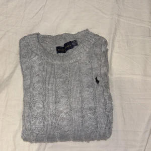 Grå kabelstickad tröja Ralph Lauren - Kabelstickad grå tröja från Ralph Lauren i bomull. Klassisk rund halsringning och långärmad modell med broderad logga på bröstet. Perfekt för dig som gillar en stilren och tidlös look. Har bara blivit Andvänd 3 gånger 