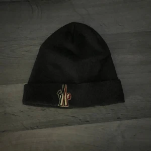 Svart Moncler mössa i ull - Svart ribbstickad mössa från Moncler med uppvikt kant och klassisk Moncler-logga framtill. Tillverkad i mjuk ull som håller dig varm under kyliga dagar. Perfekt accessoar för en stilren och exklusiv look.