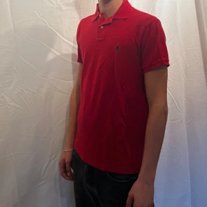 pikétröja från Ralph Lauren - Klassisk röd pikétröja från  Ralph Lauren med broderad logga på bröstet. Tröjan har krage, två knappar och korta ärmar. Tillverkad i bomull och har en slim fit passform. Perfekt för en stilren och sportig look. Modellen är 180cm.