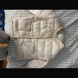 Beige quiltad dunjacka med knappar från Gina Tricot - Quiltad beige dunväst från Gina Tricot med snygga virkade kanter och två fickor framtill. Västen har rund halsringning och stängs med knappar. Perfekt för lager-på-lager och ger en clean look till din outfit.