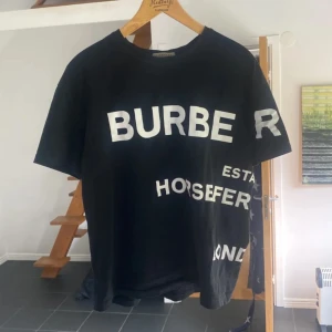 Svart Burberry t-shirt  - Svart t-shirt från Burberry med stor vit texttryck både fram och bak. Klassisk rund hals och korta ärmar. Materialet är mjuk bomull som känns skönt mot huden. Snygg streetstil med coolt Burberry London England-tryck.  Storlek L. Skick 8/10